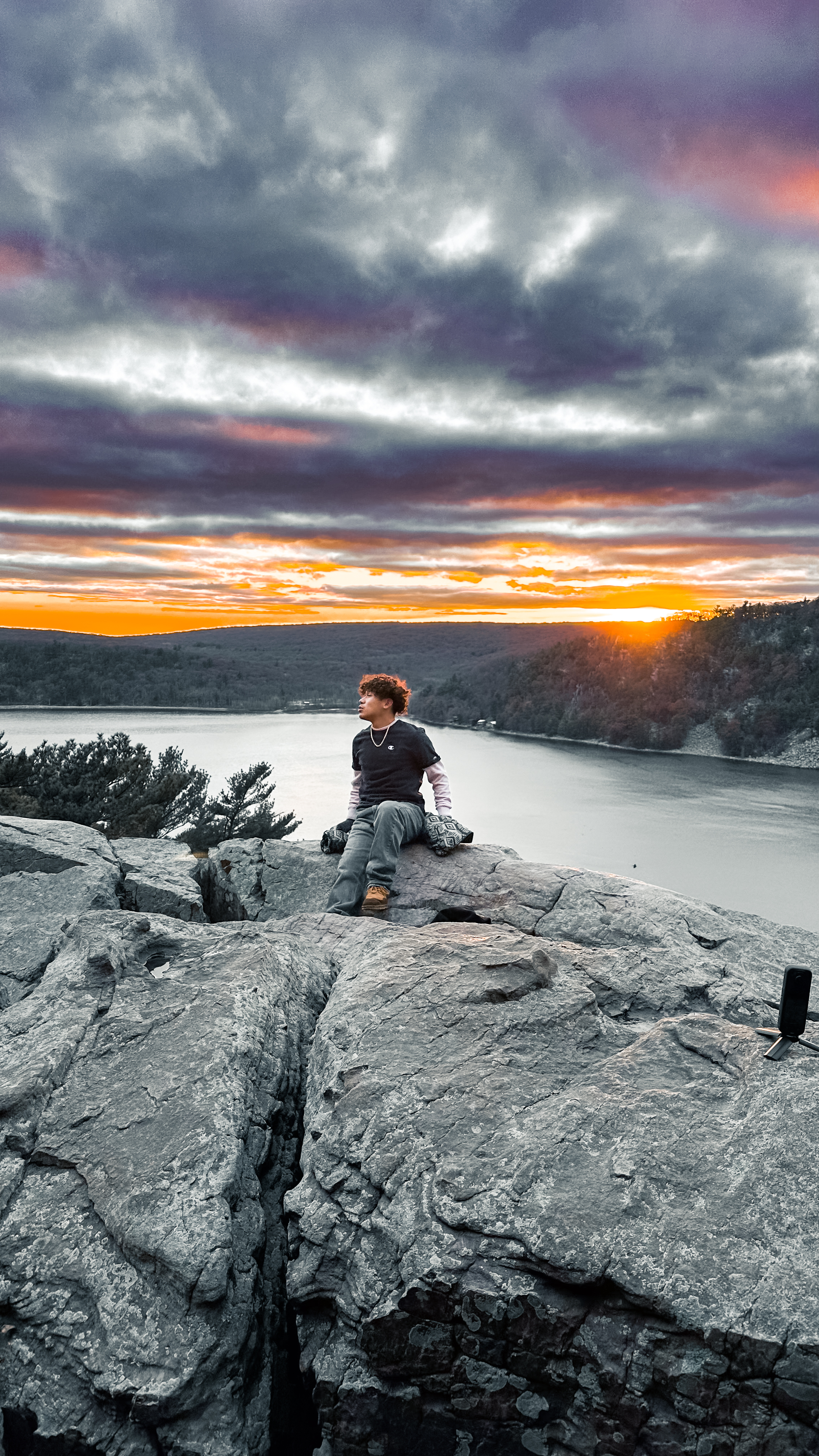 Person seated on cliff above lake with dramatic sunset sky